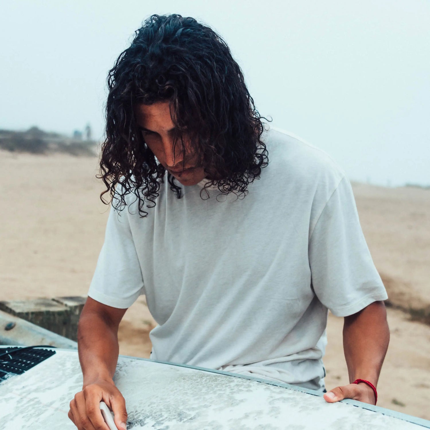 A man with long, wavy hair and his surfboard by the sea