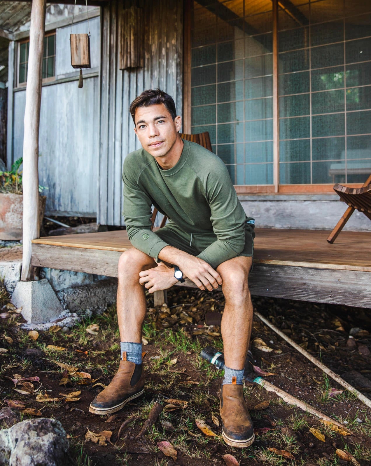 A guy sitting in front of a wooden house, wearing our alpaca Terry garments in green