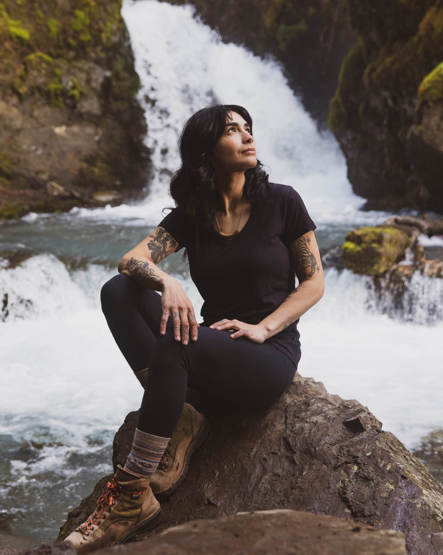 A lady in her black alpaca v-neck sitting in a rock by a river and a waterfall