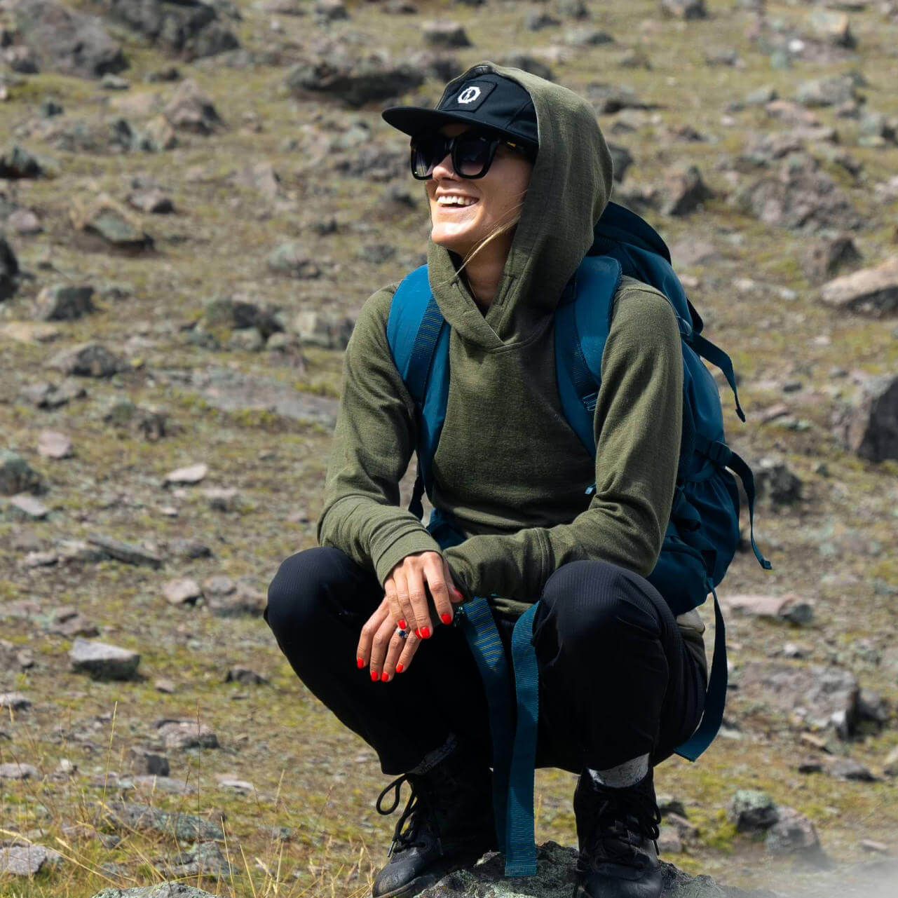 A smiling woman crouched down wearing a green Women's Hoodie and a Paka's Team 5-panel hat