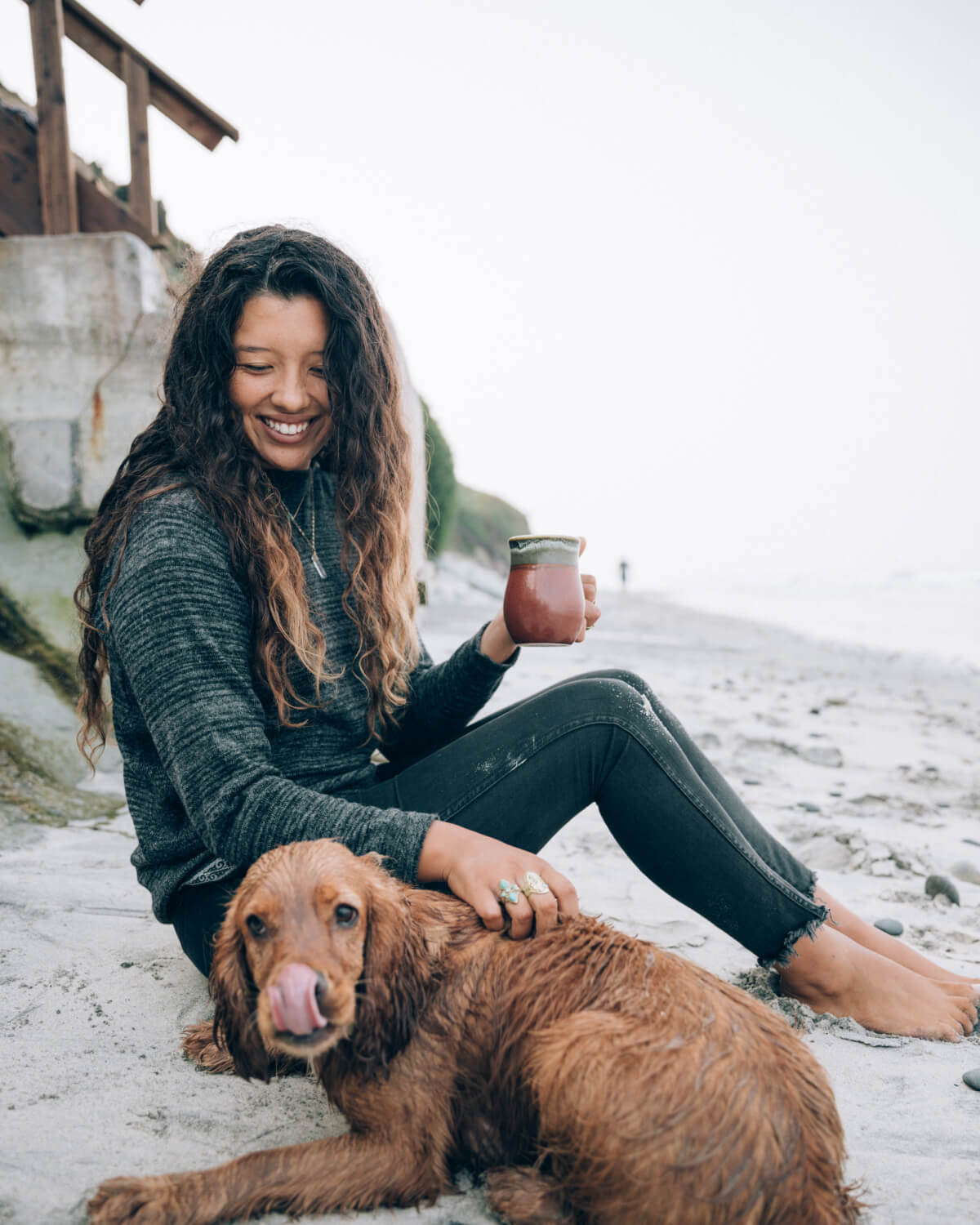 A woman petting her dog and holding a cup in a beach