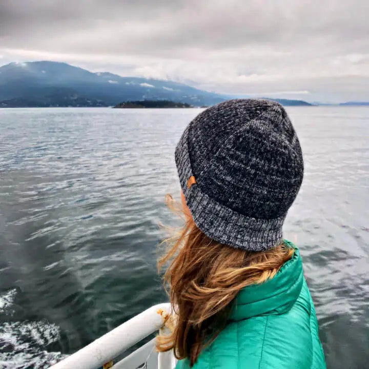 A woman sailing looking at the ocean. She's wearing al alpaca beanie