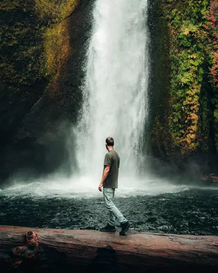 Man walking across fallen tree in front of waterfall wearing alpaca tee
