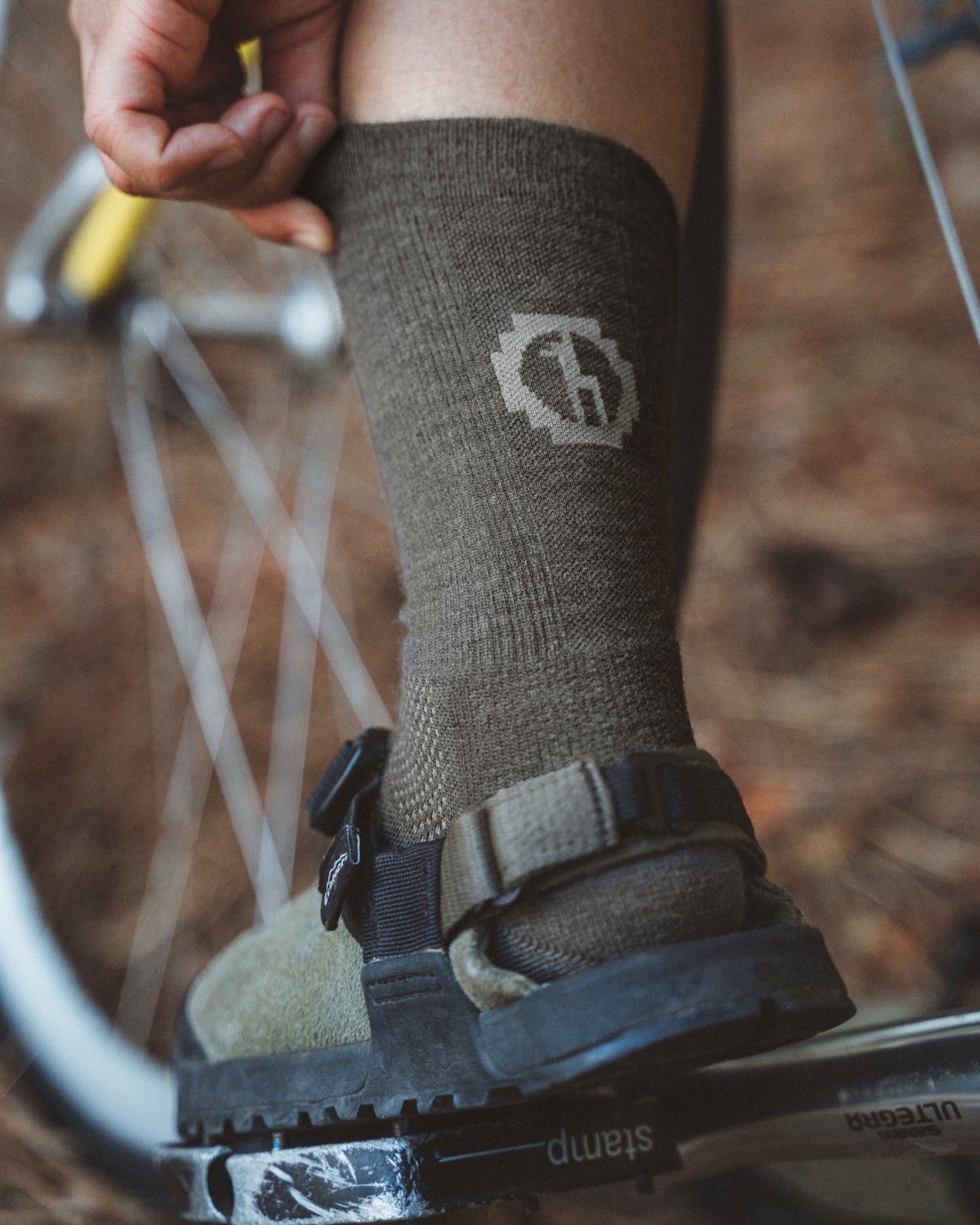 Side view of Trail Crew Socks in forage green on a model showing mid-calf crew height, ribbed texture and logo with sandal and bike-pedal background