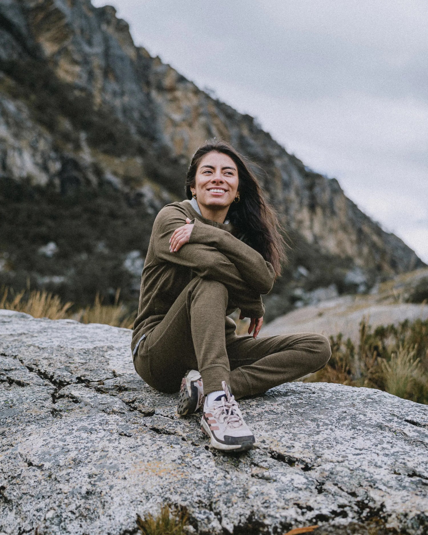 Woman sitting on a rock wearing Andean Moss women’s Breathe alpaca joggers, smiling in a mountain outdoor setting