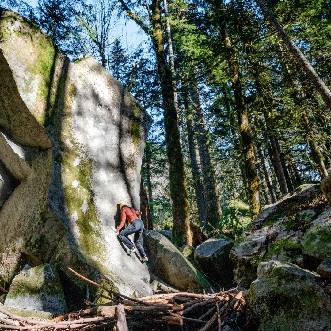 Woman in clay baselayer climbing a rock. There are some trees in the background