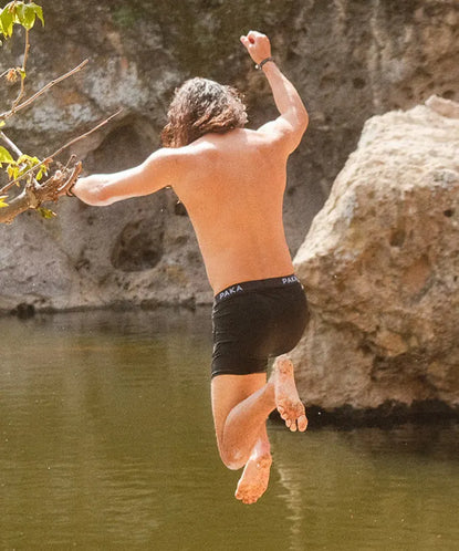 Shirtless man jumping off a rock into a lake while wearing black swim shorts