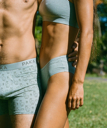 Two models wearing light gray alpaca-blend underwear, boxer briefs and high-cut briefs, standing outdoors among greenery