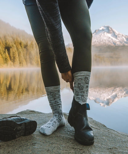 Person wearing black leggings and patterned wool socks standing on a rock by a mountain lake reflecting snow-capped peaks