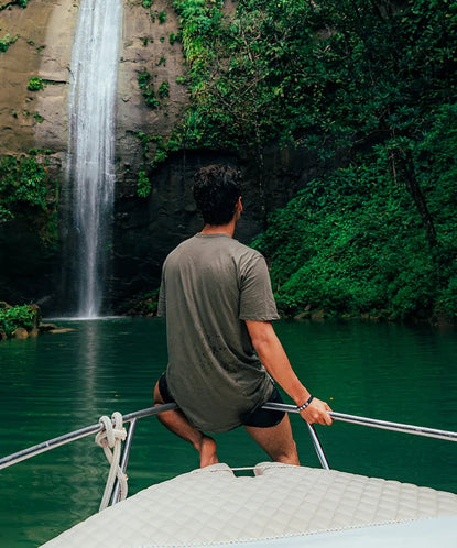 Man sitting on the bow of a boat looking toward a waterfall surrounded by lush green cliffs