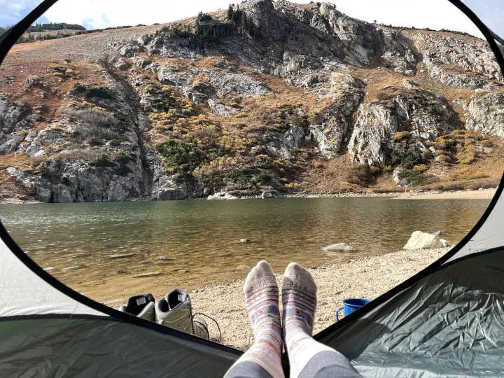 A picture of feet wearing our Inca socks taken from a tent with a great view of the mountains and a lake