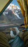 A man wearing our light grey socks in a tent outdoors. There is a snowy mountain in the background