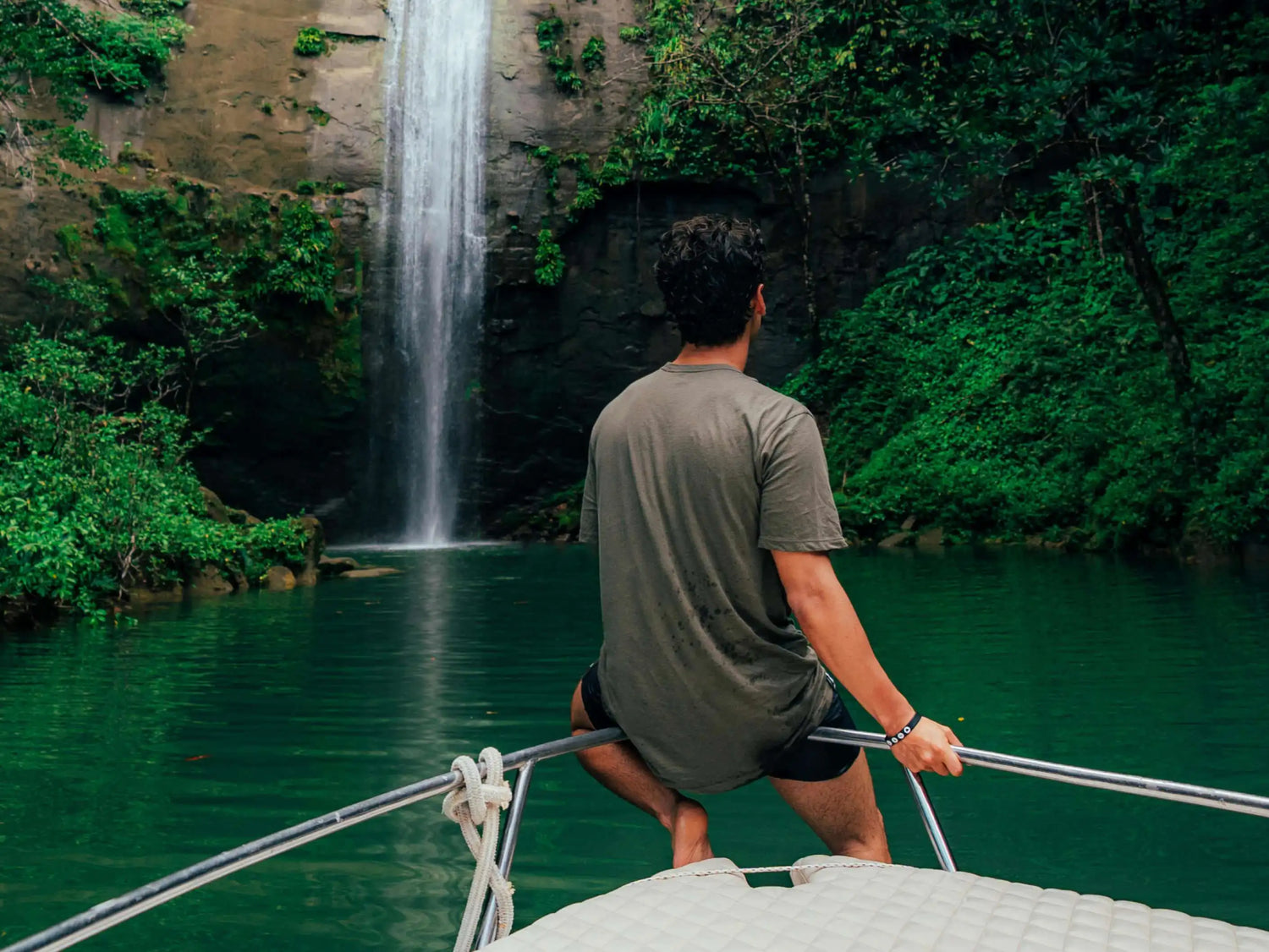 A man in a boat wearing a green alpaca tee in front of a beautiful waterfall and lake 