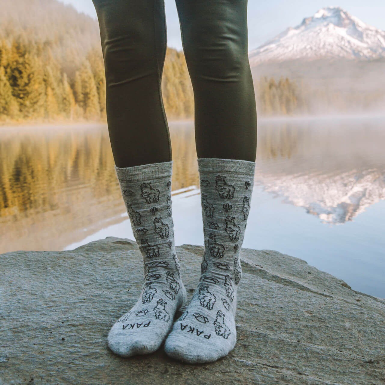 Persons feet in alpaca socks standing in front of lake with the reflection of a mountain