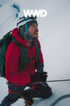 A man wearing a red Pakafill puffer jacket and a helmet summitting a snowy mountain