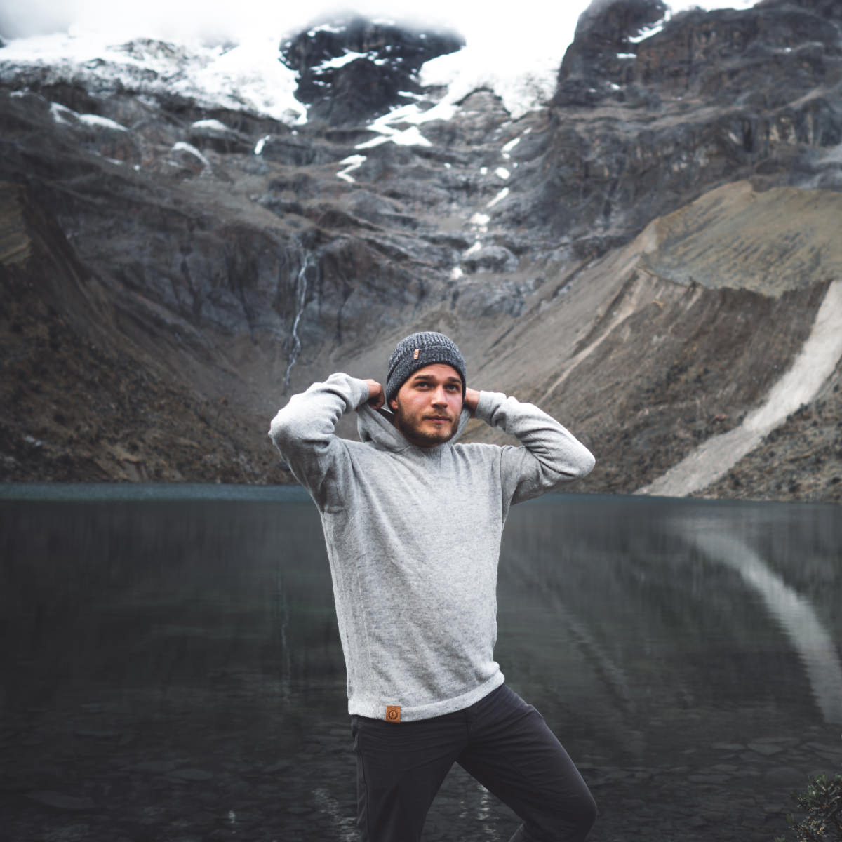 A man wearing a light grey alpaca hoodie in front of a lake and a snowy mountain