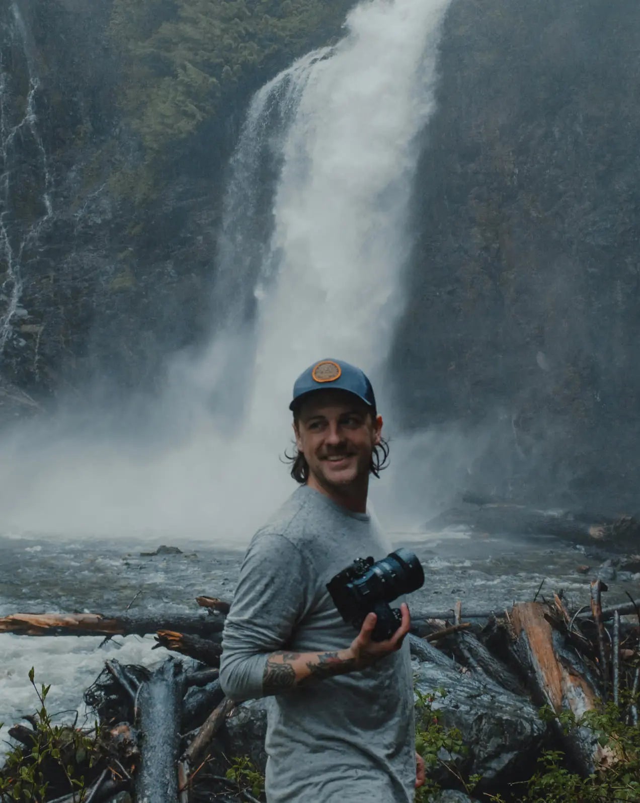 A smiling man holding a camera with a beautiful waterfall behind him
