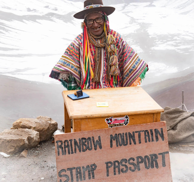 A man wearing a colorful poncho. With a stand and a sign that says "Rainbow Mountain Stamp Passport"