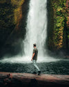 Man walking across fallen tree wearing green alpaca tee in front of waterfall 
