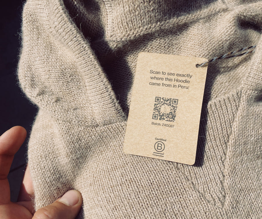 A light grey alpaca hoodie with a tag that says "scan to see exactly where this Hoodie came from in Peru" It also includes a QR code 