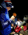 Man with headlamp and flashlight in the dark with two other people making tea. They are all bundled up in coats and hats. 