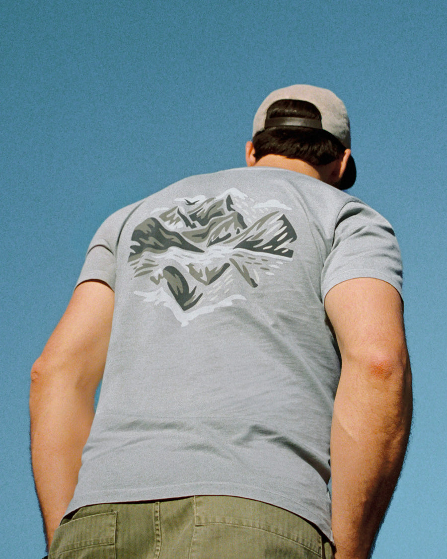 Person wearing a gray PAKA "Love Nature Back" t-shirt with a graphic design on the back against a clear blue sky.