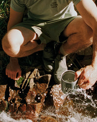 Person squatting by a stream, washing something in a cup.