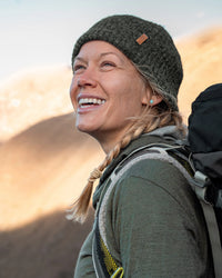 Smiling woman hiker wearing a dark green marled beanie and backpack in a sunlit mountain landscape
