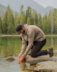 Man kneeling at a mountain lake scooping water while wearing a timber-colored slim-fit alpaca full-zip hoodie