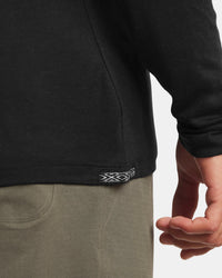 Close-up view of Men’s Aire long sleeve alpaca shirt showing hem seam and woven tab with model’s hand and olive pants