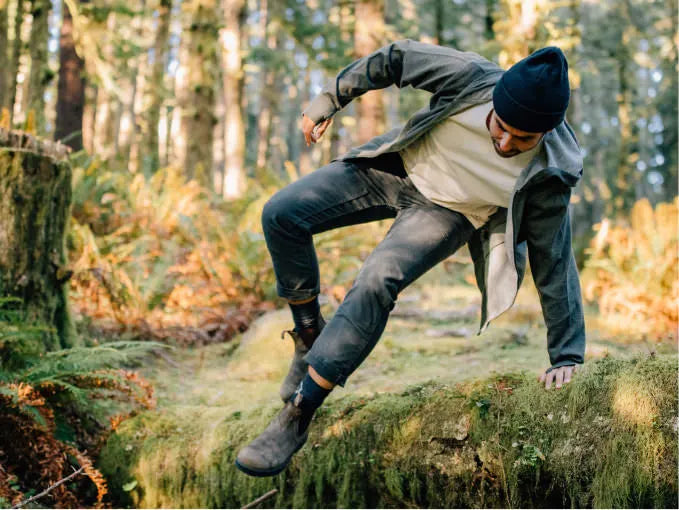 A man wearing a green full zip hoodie jumping over a tree trunk in the nature