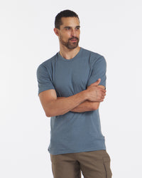 Front view of men’s tee in blue on model with arms crossed showcasing short sleeves and lightweight fit on white background