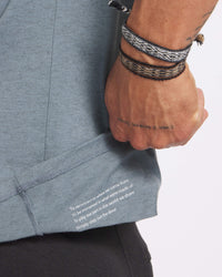 Close-up of Men’s Tee hem in steel showing printed text ‘To reconnect to where we come from’ with hand holding hem