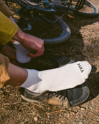 Side view of white Trail 1/4 crew socks on a person showing low crew height and PAKA logo on the toe, dirt trail with bike