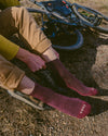 Model pulling on maroon 3/4 crew PAKA hiking socks showing PAKA logo on toes, sitting by a gravel trail with a mountain bike