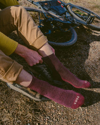 Model pulling on maroon 3/4 crew PAKA hiking socks showing PAKA logo on toes, sitting by a gravel trail with a mountain bike