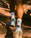 Rear view of Trail 3/4 Crew socks in white showcasing 3/4 crew height and PAKA cuff logo on a model walking on a rocky trail