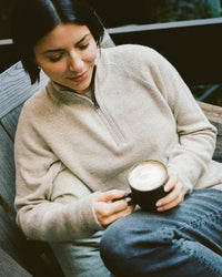 Model wearing pebble alpaca quarter-zip sweater in a regular fit, seated holding a mug on a bench outdoors