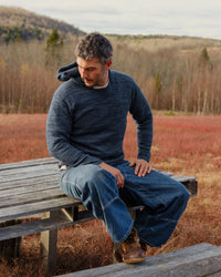 Seated three-quarter view of The Hoodie alpaca hoodie in navy showing marled knit texture, hood, and relaxed fit on a model outdoors