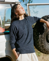 Model wearing navy alpaca hoodie standing beside a vehicle with a spare tire, hands in pockets and face turned toward the sun