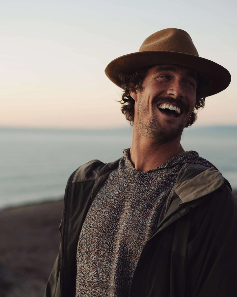 Man wearing a brown hat and jacket standing on a beach with ocean in the background