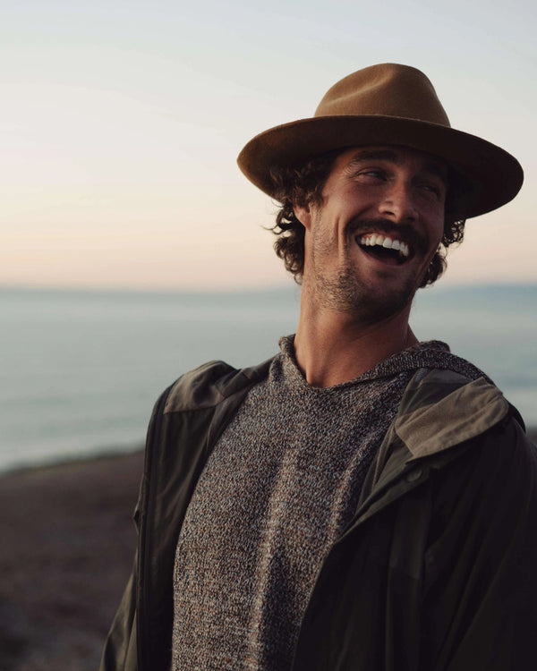 Man wearing a brown hat and jacket standing on a beach with ocean in the background