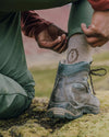 Person pulling up a PAKA Trail 3/4 crew hiking sock in light tan while wearing a muddy hiking boot on mossy ground