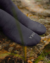 Close-up of black Trail 3/4 crew socks on feet showing PAKA logo on toes and textured knit, resting on a rocky trail