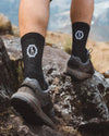 Back view of black Trail 3/4 crew socks on model showing mid-calf coverage and white logo with rocky hiking trail background