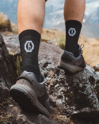 Back view of black Trail 3/4 crew socks on model showing mid-calf coverage and white logo with rocky hiking trail background