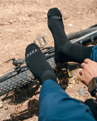 Person wearing black Trail 3/4 crew socks with PAKA on the toes, feet resting on a mountain bike tire on a rocky trail