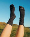Legs wearing charcoal mid-calf Trail Crew alpaca socks raised over grassy hills under blue sky