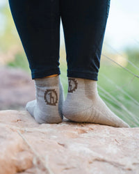 Rear view of charcoal blue Trail 1/4 Crew socks on a model showing low-crew height, heel logo, and cushioned heel on rocky outdoor background