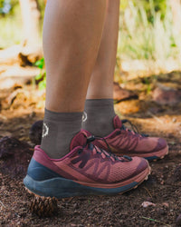 Side view of Trail 1/4 Crew lightweight hiking socks in charcoal with white logo, worn on model on a rocky trail